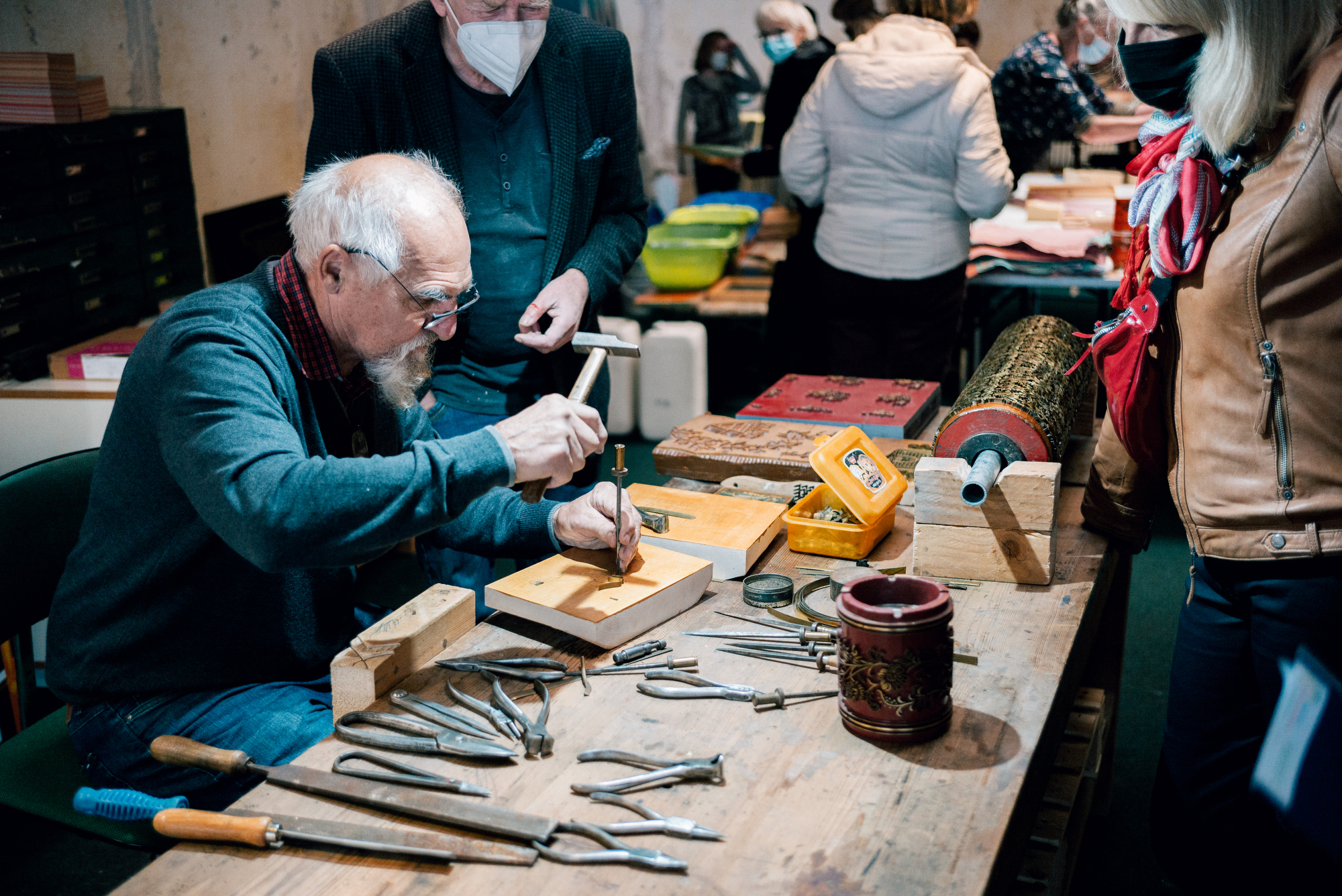 Ein Mann prägt mit Hammer und Werkzeug Metallplatten, umgeben von Zuschauern in einer Werkstatt.